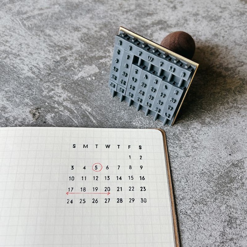 May include: A close-up of a calendar with the number 5 circled in red. The calendar is on a gray surface. The date is the 5th of the month.