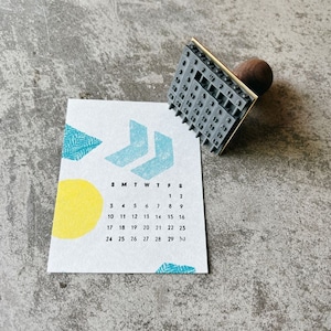 May include: A gray rubber stamp with a calendar design. The stamp is mounted on a wooden handle and has a gold metal frame. The calendar shows the days of the week and the numbers 1 through 30.