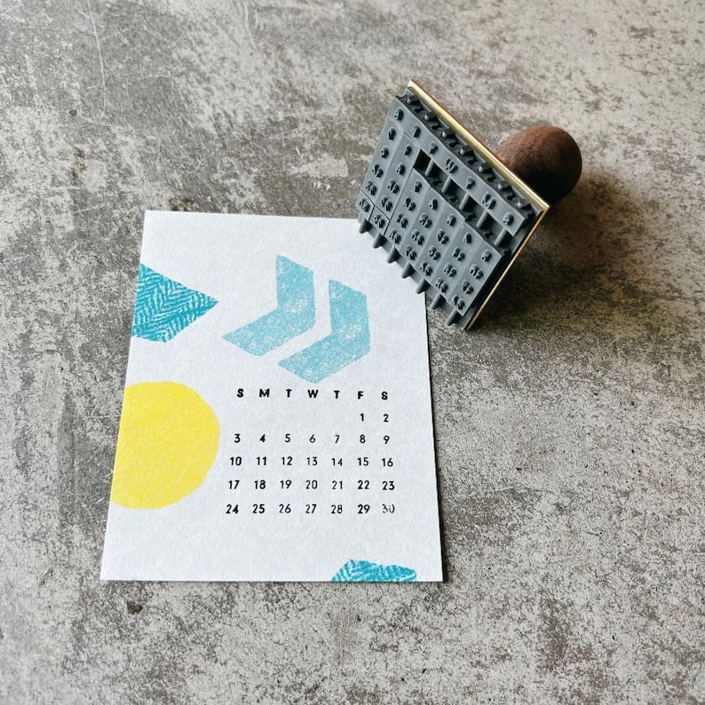 May include: A gray rubber stamp with a calendar design. The stamp is mounted on a wooden handle and has a gold metal frame. The calendar shows the days of the week and the numbers 1 through 30.