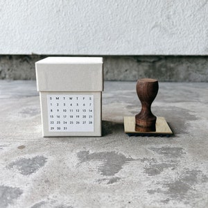May include: A white box with a calendar printed on the front. The calendar shows the days of the week and the numbers 1 through 31. A wooden stamp with a brass base is sitting on a gray surface.
