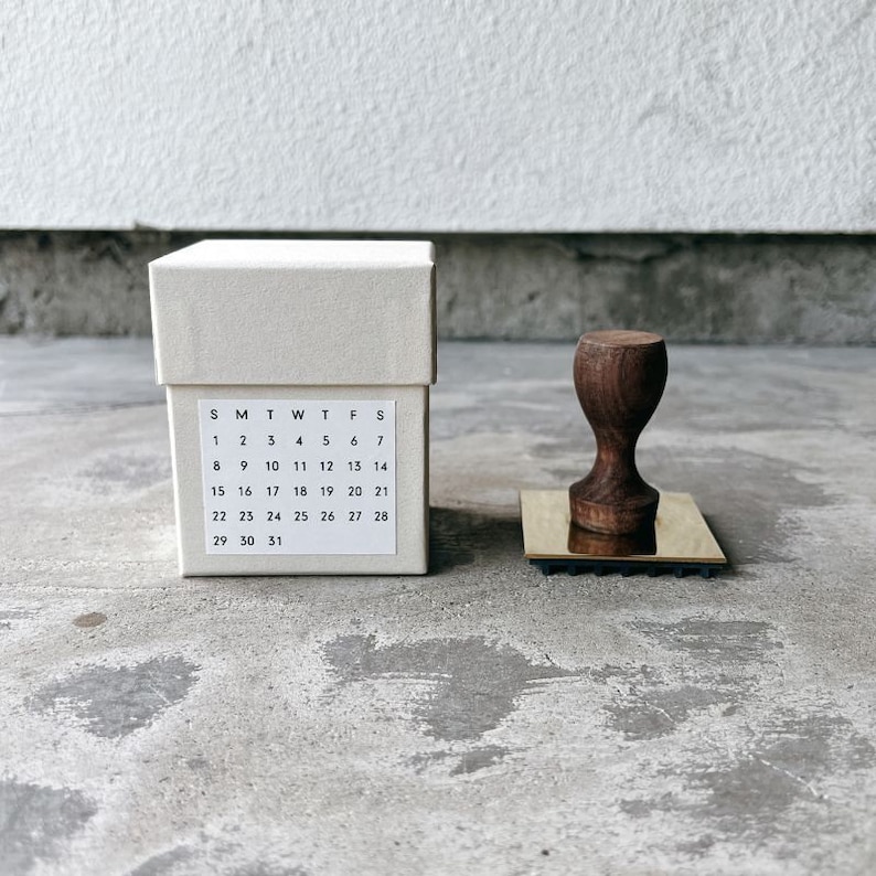 May include: A white box with a calendar printed on the front. The calendar shows the days of the week and the numbers 1 through 31. A wooden stamp with a brass base is sitting on a gray surface.
