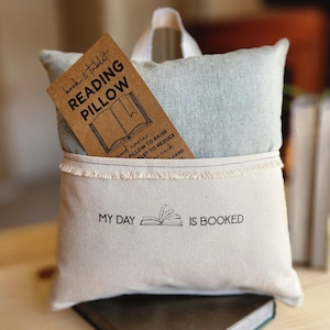 May include: A beige canvas pillow with a pocket that holds a brown card with the text "book & tablet reading pillow". The pillow has a frayed edge and the text "My day is booked" printed on the front.