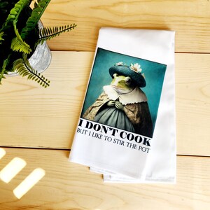May include: White kitchen towel with a green frog wearing a blue hat and a white lace collar. The text on the towel reads "I don't cook but I like to stir the pot."