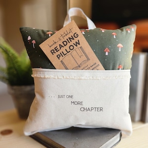 Op de afbeelding: Een groen en wit kussen met een paddenstoelprint en een zakje aan de voorkant. Het zakje heeft een bruine kaart met de tekst "book & tablet reading pillow" en de tekst "... just one more chapter" is op het zakje gedrukt.