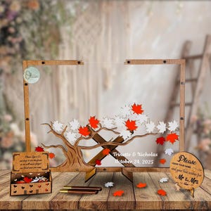 Puede incluir: Un libro de visitas alternativo de madera con un diseño de árbol, con recortes de hojas de arce rojas y blancas. El marco incluye los nombres "Trinette & Nicholas" y la fecha "October 24, 2025". Incluye una pequeña caja y un letrero que dice "Please leave your name and your best wishes for Mr & Mrs".
