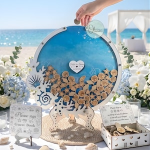 Op de afbeelding: Een ronde, witte houten gastenboek-alternatief met een blauwe oceaan-thema achtergrond. Het frame is versierd met houten zeesterren en schelpen. Inclusief een bord met de tekst "Please sign my Guest Book". Een hand laat een houten schelp in het frame vallen.