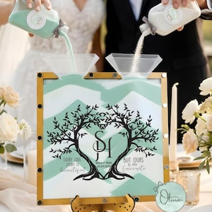 May include: A wedding sand ceremony set with a gold-framed display. The display features a tree design with initials and the text "Every love story is beautiful, but ours is my favourite." Two people pour sand into the display.
