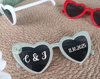 May include: Heart-shaped sunglasses in white, red, and light green. The light green pair has the initials "e & J" and the date "11.10.2025" printed on the lenses. The sunglasses are arranged on a textured, neutral-toned surface.