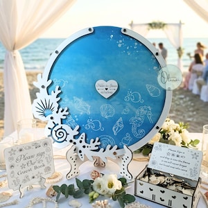 Könnte beinhalten: Ein runder, blauer Gästebuch-Ersatz mit weißem Rahmen und korallenförmigen Akzenten. Die blaue Mitte zeigt Muschelbilder und die Namen "Helen & Steven" mit einem Datum. Zusätzliche Schilder und eine Box für Gästesignaturen sind ebenfalls vorhanden.