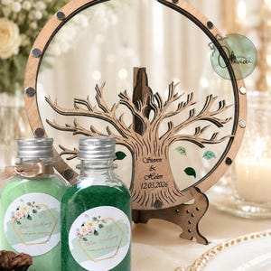 May include: A wooden wedding guest book alternative with a tree design, featuring the names "Steven & Helen" and the date "12.03.2026". Two small glass bottles filled with green and light green sand are in front of the guest book.
