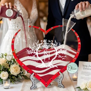 Può includere: Un set per la cerimonia della sabbia dell'unità a forma di cuore con sabbia rossa e bianca. La cornice è rossa con dettagli argentati e un disegno ad albero. La cerimonia si svolge a un matrimonio.