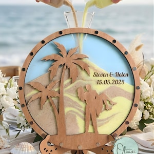 Puede incluir: Una pantalla de ceremonia de arena de madera con un tema de playa. La pantalla presenta un marco circular con un diseño de palmera y silueta de pareja. Los nombres "Steven & Helen" y la fecha "15.05.2025" son visibles. Dos botellas vierten arena en la pantalla.