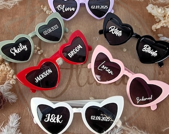 May include: A collection of heart-shaped sunglasses in various colors, including lavender, green, red, black, pink, and white. Each pair features personalized text such as names and dates, perfect for a wedding or special event.