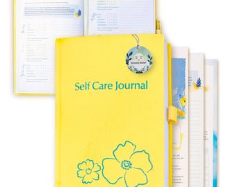 May include: Yellow self-care journal with a teal floral design and the text "Self Care Journal". A teal circle with a moon and the text "Anxiety Relief" is attached to the journal.