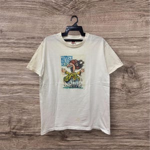 Puede incluir: Camiseta blanquecina con un dibujo animado colorido y el texto "Hippies Delight". El dibujo representa un personaje con pelo alborotado y una amplia sonrisa. La camiseta está colgada de una percha negra.