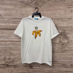 May include: White t-shirt featuring a hand-drawn banana design. The banana is yellow with brown outlines and a green crown. The shirt is hanging on a black hanger against a wooden background.