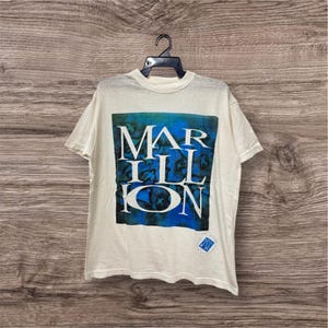 以下が含まれることがあります： 白に近い色のTシャツ。バンド名「MARILLION」が白の英大文字で、青と緑のグラフィックの上に配置されています。Tシャツはクルーネックと半袖です。小さな青いダイヤモンドのロゴが見えます。