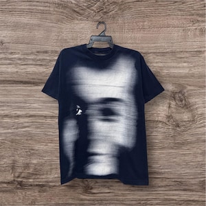 以下が含まれることがあります： 濃いネイビーブルーの半袖Tシャツで、中央に人間の顔が水平方向にぼやけた大きな白いグラフィックプリントが施されています。胸の中央を覆うデザインで、動きのある歪んだ肖像画と目元のディテールが特徴です。