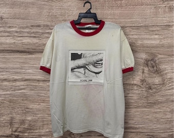May include: A cream-colored ringer t-shirt with red trim on the collar and sleeve cuffs. The front features a rectangular black and white graphic print of a mouth and a bandaged object, with the text PEARL JAM printed below the image.