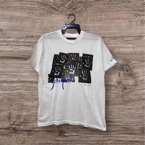 Puede incluir: Camiseta blanca con un diseño gráfico que presenta múltiples cuadrados negros, cada uno con una huella de mano blanca. La palabra "chimme" está escrita en escritura azul debajo de los cuadrados. La camiseta está colgada de una percha negra.