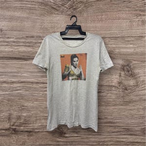 May include: Light gray t-shirt featuring a graphic of a woman in a boxing pose. The image has a vintage aesthetic with a color palette of orange, yellow, and brown. The t-shirt is hanging on a black hanger.