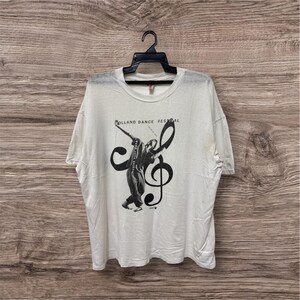 Puede incluir: Camiseta blanca con un gráfico negro de un bailarín y una clave de sol. El texto "HOLLAND DANCE FESTIVAL" está encima del bailarín. La camiseta está colgada de una percha negra.