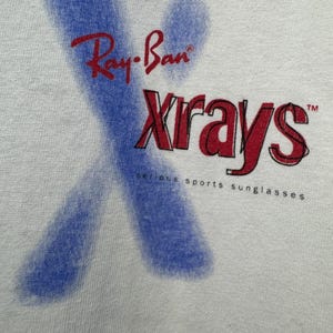 May include: White t-shirt featuring the Ray-Ban logo in red, with the word "Xrays" in red and black, and a blue X-shaped design. The text "serious sports sunglasses" is printed below the logo.