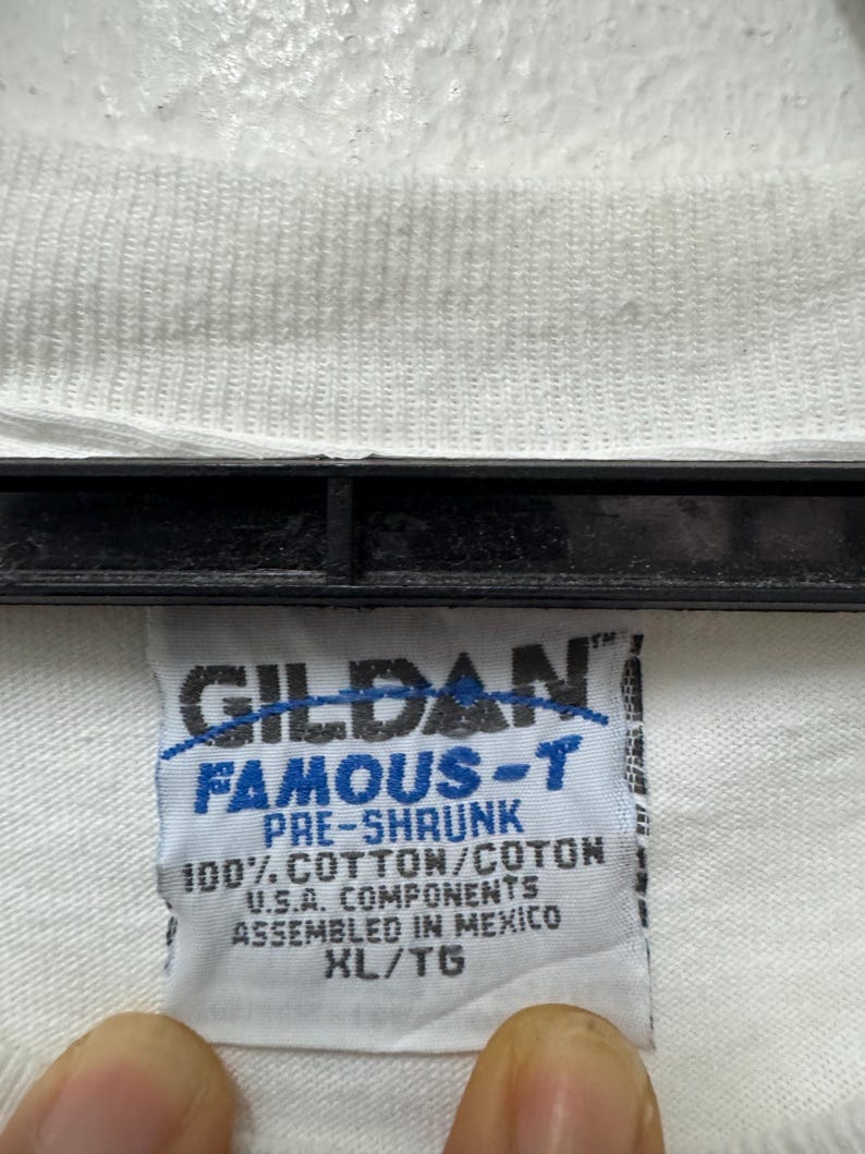 May include: Close-up of a white t-shirt tag with the brand name "GILDAN FAMOUS-T" printed in blue and black. The tag also includes text indicating the shirt is pre-shrunk, made of 100% cotton, and assembled in Mexico. The size is XL/TG.