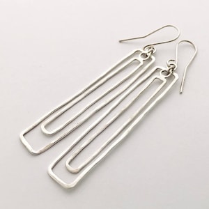 Könnte beinhalten: Ein Paar silberne Ohrhänger mit geometrischem Design. Die Ohrringe bestehen aus mehreren dünnen, rechteckigen Metallteilen, die übereinander gelegt sind.