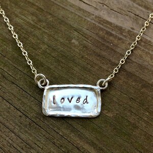 May include: A silver necklace with a rectangular pendant that says "loved" on it. The pendant has a hammered texture.