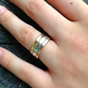 May include: Three stacked hammered metal rings in silver, gold, and rose gold. The rings are worn on a person's finger.