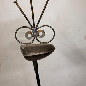 May include: A whimsical metal sculpture resembling a face. It features circular eyes, three nail antennae, and a rounded head. The sculpture is mounted on a black rod, creating a unique and artistic piece.