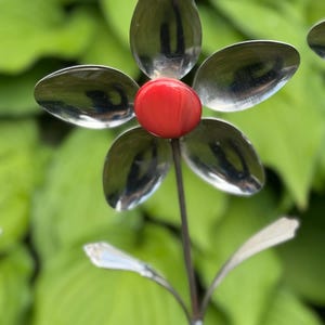 May include: A handcrafted metal flower sculpture with spoon-shaped petals and a red center. The flower is mounted on a slender stem with two leaf-shaped accents. The background is a soft focus of green foliage.