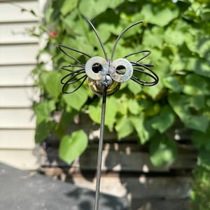 May include: A whimsical metal garden art piece depicting a stylized insect. The insect features a brass head, metal washers for eyes, and wire antennae and wings. The sculpture is mounted on a slender metal rod, perfect for outdoor display.