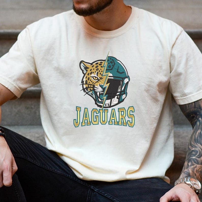 Puede incluir: Una camiseta de manga corta color crema con un gr&aacute;fico de una cabeza de jaguar dividida por un rayo junto a un casco de f&uacute;tbol americano color verde azulado. La palabra JAGUARS aparece en letras amarillas con borde verde azulado.