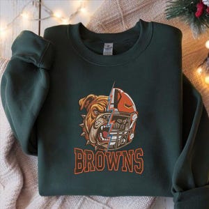 Puede incluir: Una sudadera de cuello redondo verde oscuro con un estampado gr&aacute;fico de una cara de bulldog dividida con un casco de f&uacute;tbol americano. Un rayo azul separa las dos mitades. La palabra BROWNS est&aacute; impresa en letras naranjas debajo del dise&ntilde;o.