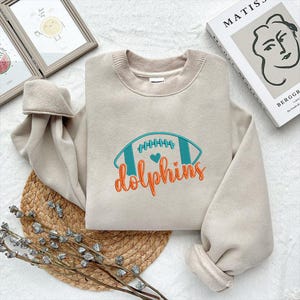 Puede incluir: Una sudadera beige con un dise&ntilde;o de f&uacute;tbol bordado en verde azulado y naranja, con la palabra "dolphins" debajo. La sudadera tiene cuello redondo y mangas largas con pu&ntilde;os acanalados. El dise&ntilde;o incluye un coraz&oacute;n.