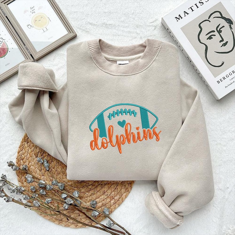 Puede incluir: Una sudadera beige con un dise&ntilde;o de f&uacute;tbol bordado en verde azulado y naranja, con la palabra "dolphins" debajo. La sudadera tiene cuello redondo y mangas largas con pu&ntilde;os acanalados. El dise&ntilde;o incluye un coraz&oacute;n.