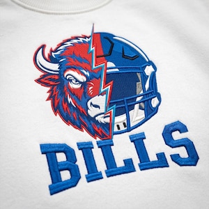 Puede incluir: Una sudadera blanca con un bordado que muestra una cabeza de bisonte dividida por un rayo y un casco de f&uacute;tbol americano. La palabra BILLS est&aacute; bordada en letras azules de bloque debajo del gr&aacute;fico, creando un dise&ntilde;o texturizado y llamativo.