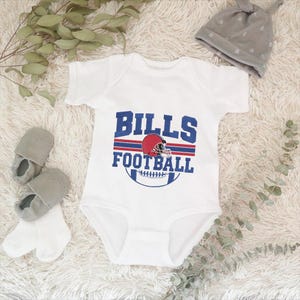 May include: White baby bodysuit with "BILLS FOOTBALL" in blue and red lettering, featuring a football helmet graphic. Includes gray baby shoes, white socks, and a gray beanie with a knotted top.