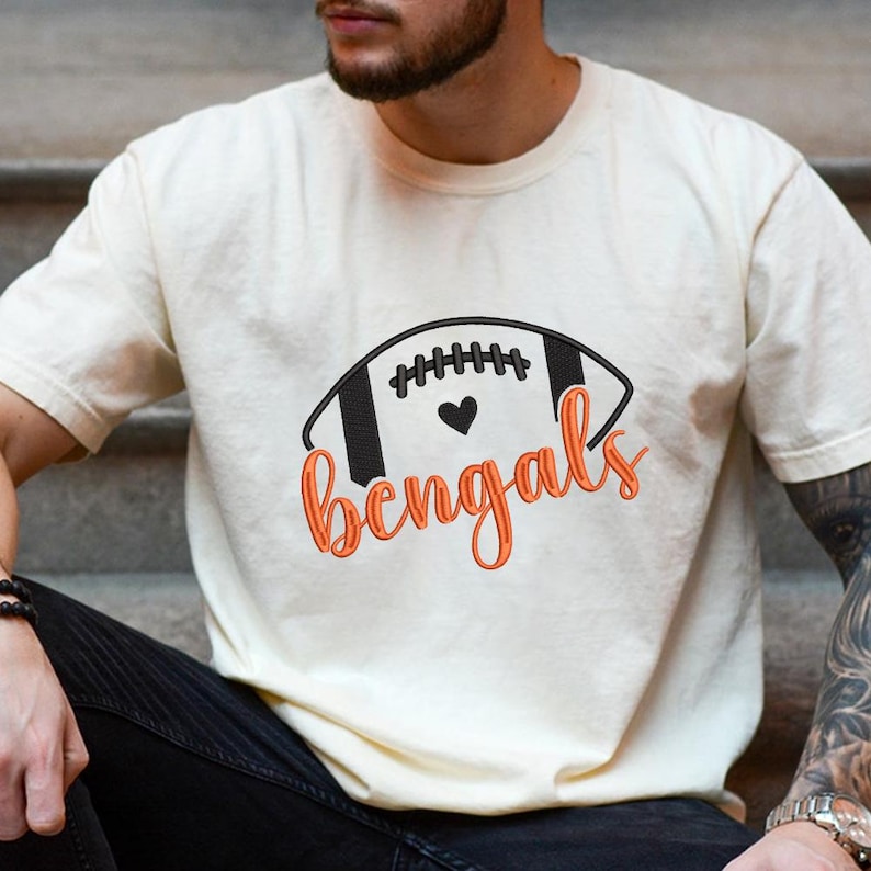 Puede incluir: Camiseta color crema con un dise&ntilde;o de f&uacute;tbol americano y la palabra "Bengals" en escritura naranja. El gr&aacute;fico del bal&oacute;n de f&uacute;tbol es en blanco y negro con un coraz&oacute;n en el centro. Una prenda de vestir casual y deportiva.
