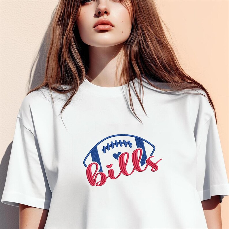 May include: White t-shirt featuring a blue and red football design with the word "Bills" in a script font. The football has blue stripes and white stitching. Two small hearts are inside the football.