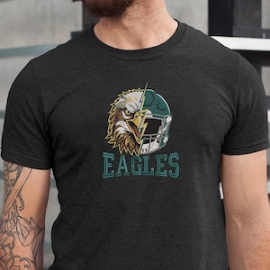 Puede incluir: Una camiseta gris carb&oacute;n de manga corta con un dise&ntilde;o gr&aacute;fico que divide la cabeza de un &aacute;guila calva y un casco de f&uacute;tbol americano verde oscuro con un rayo. La palabra EAGLES est&aacute; impresa en letras de bloque color verde azulado.