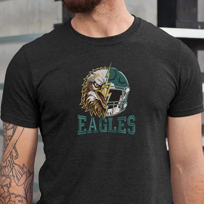 Puede incluir: Una camiseta gris carb&oacute;n de manga corta con un dise&ntilde;o gr&aacute;fico que divide la cabeza de un &aacute;guila calva y un casco de f&uacute;tbol americano verde oscuro con un rayo. La palabra EAGLES est&aacute; impresa en letras de bloque color verde azulado.