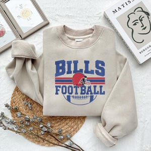 May include: Beige sweatshirt with the text "BILLS FOOTBALL" in blue, red, and white. The design includes a football helmet and a football. The sweatshirt is folded on a woven mat.