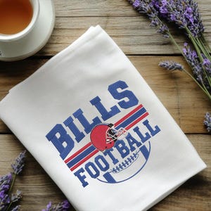 May include: White folded towel with the words "BILLS FOOTBALL" in blue and red. A red football helmet is in the center of the design. The towel is on a wooden surface with a cup of tea and lavender sprigs.