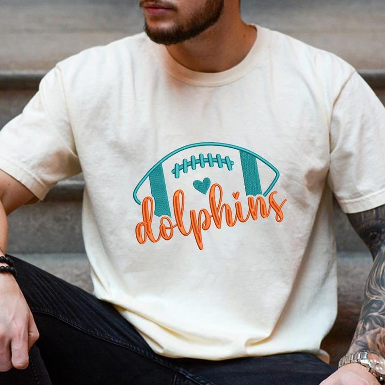 Puede incluir: Camiseta color crema con un dise&ntilde;o de f&uacute;tbol americano turquesa y naranja con la palabra "Dolphins". El gr&aacute;fico de f&uacute;tbol incluye un coraz&oacute;n y detalles de costura. La camiseta es de un material suave.