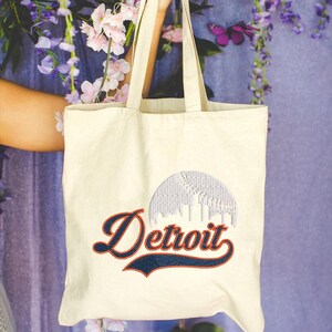 May include: Cream-colored tote bag featuring an embroidered baseball design and the word "Detroit" in a script font. The baseball design is white and the text is navy blue with an orange outline. The bag has two handles.