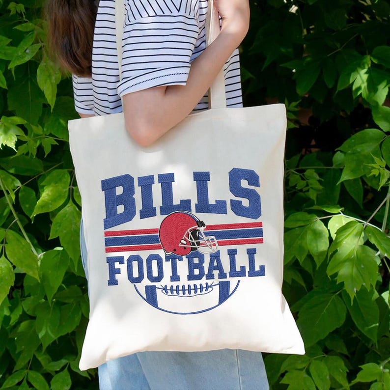 May include: Cream-colored tote bag featuring the text "BILLS FOOTBALL" in blue, with a red and blue football helmet graphic. The bag has a football graphic at the bottom. The bag is held by a person wearing a striped shirt.