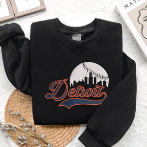 May include: Black sweatshirt with the word "Detroit" in orange script and a white baseball design. The sweatshirt has a crew neck and long sleeves. The design features a baseball with a skyline.